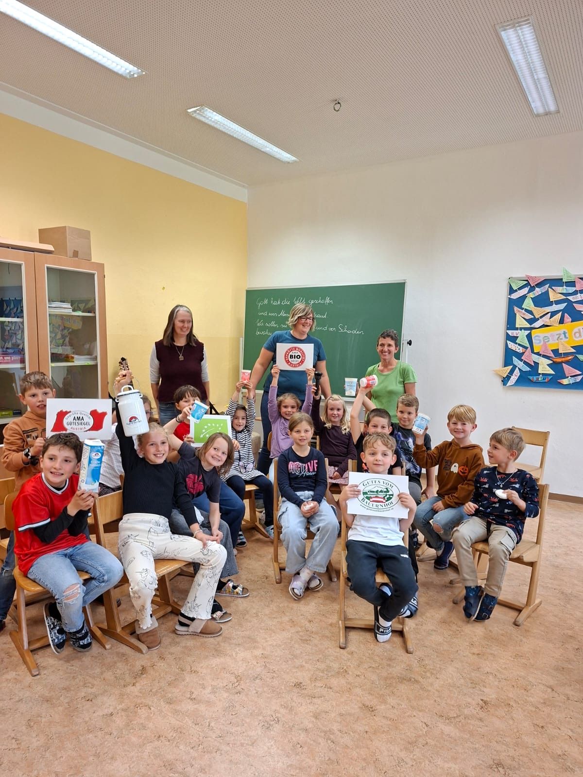 Kinder und Erwachsene in einem Klassenzimmer halten Schilder und Produkte hoch. Zwei Frauen stehen vor einer Tafel mit Schrift. Ein Bulletin Board hat farbige Papierausschnitte.