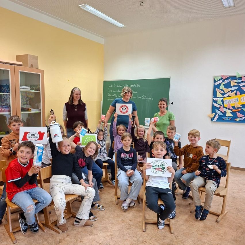 Kinder und Erwachsene in einem Klassenzimmer halten Schilder und Produkte hoch. Zwei Frauen stehen vor einer Tafel mit Schrift. Ein Bulletin Board hat farbige Papierausschnitte.