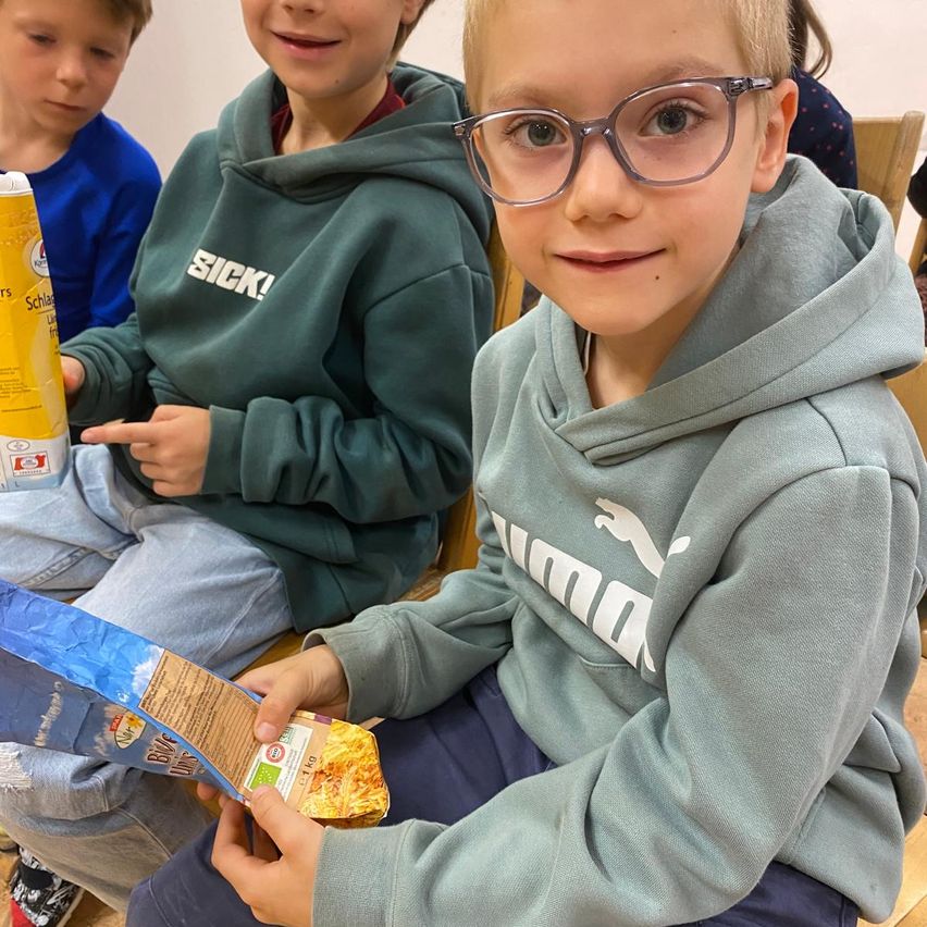 Ein junger Junge mit Brille hält eine Tüte mit Essen und lächelt, während er unter anderen sitzt, von denen zwei Kapuzenpullover tragen.