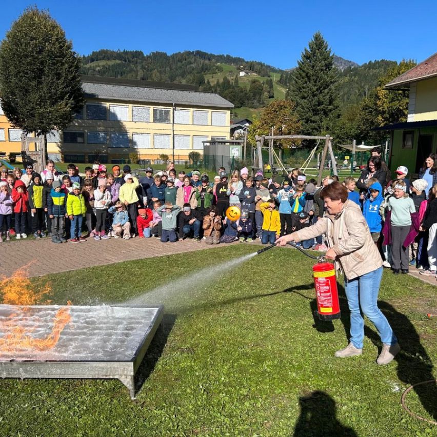 Eine Frau sprüht Wasser aus einem Feuerlöscher auf ein brennendes Objekt, während eine Menge Kinder zusieht.