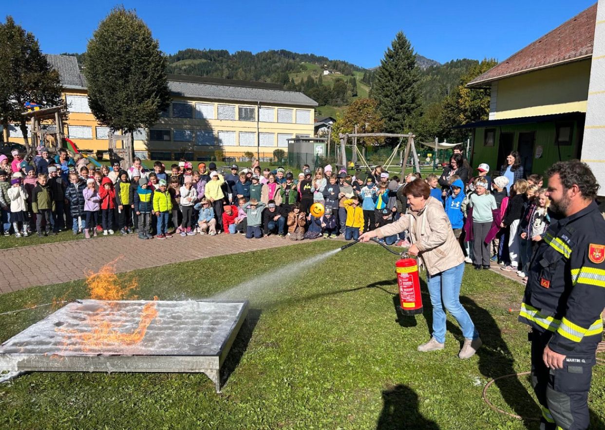 Eine Frau sprüht Wasser aus einem Feuerlöscher auf ein brennendes Objekt, während eine Menge Kinder zusieht.
