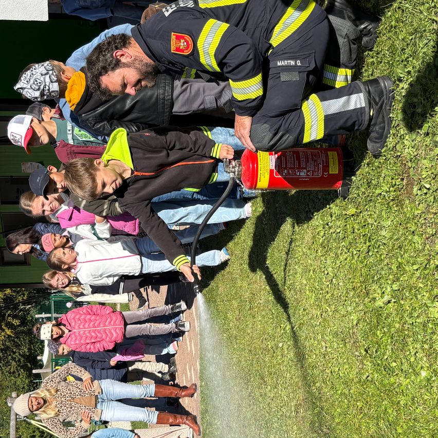 Ein Feuerwehrmann kniet sich, um Kindern in einer Reihe auf einem Rasen einen Feuerlöscher zu demonstrieren.