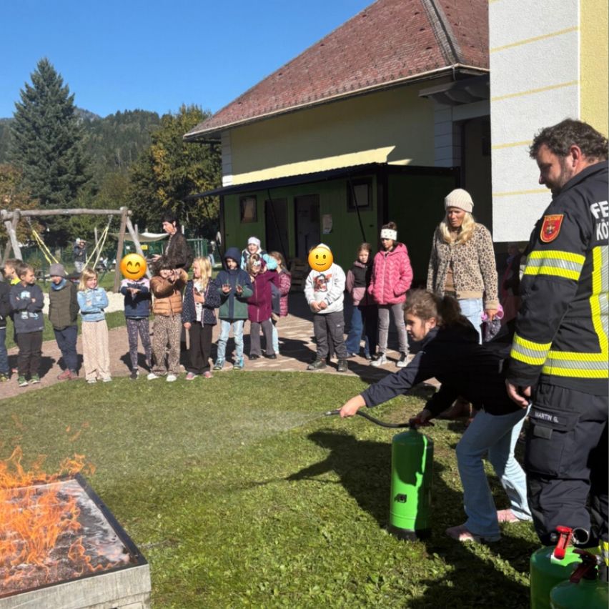 Eine Gruppe von Kindern und Erwachsenen versammelt sich vor einem Haus und beobachtet, wie ein Feuerwehrmann ein Feuer mit einem Feuerlöscher löscht. Einige tragen Winterkleidung, und zwei haben smiley-Gesichter auf ihre Gesichter gezeichnet.