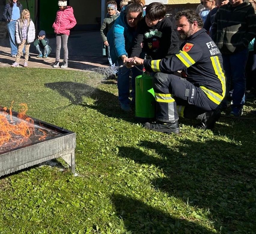 Ein Feuerwehrmann demonstriert Kindern auf einem Rasenfeld Feuerlöschtechniken, mit einem Feuerkorb in der Nähe.