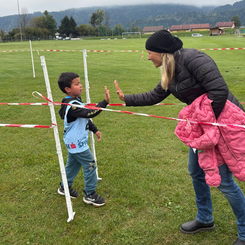 Eine Frau in einer schwarzen Jacke und Mütze begrüßt einen Jungen in einem blauen Trikot mit der Aufschrift 'SFR' auf einem Rasenfeld mit weißen Stangen und rot-weißem Band.