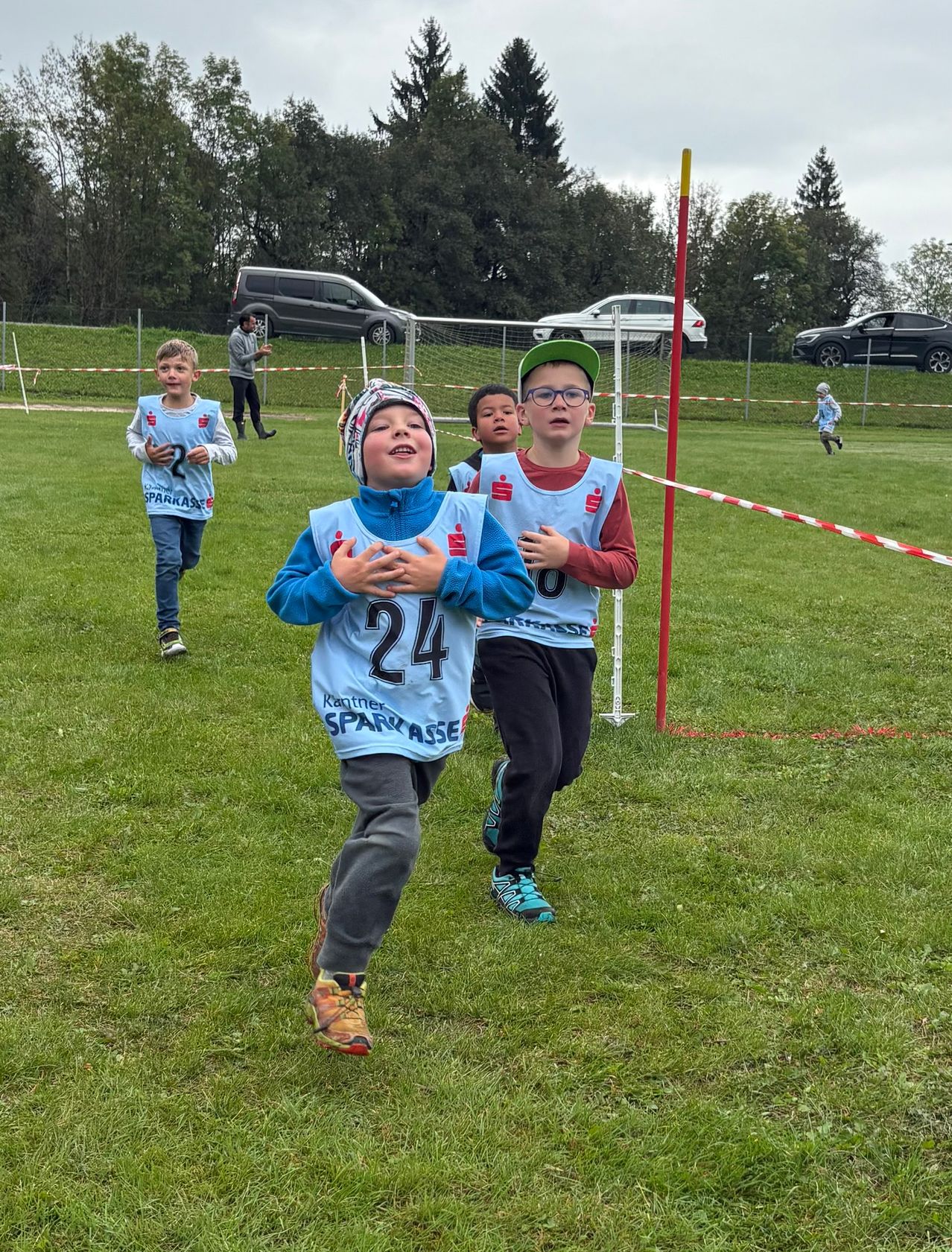 Kinder in Trikots mit Nummern laufen auf einem Rasenfeld. Sie nehmen an einem Rennen teil. Ein Fußballtor und rot-weiße Barrieren sind im Hintergrund.
