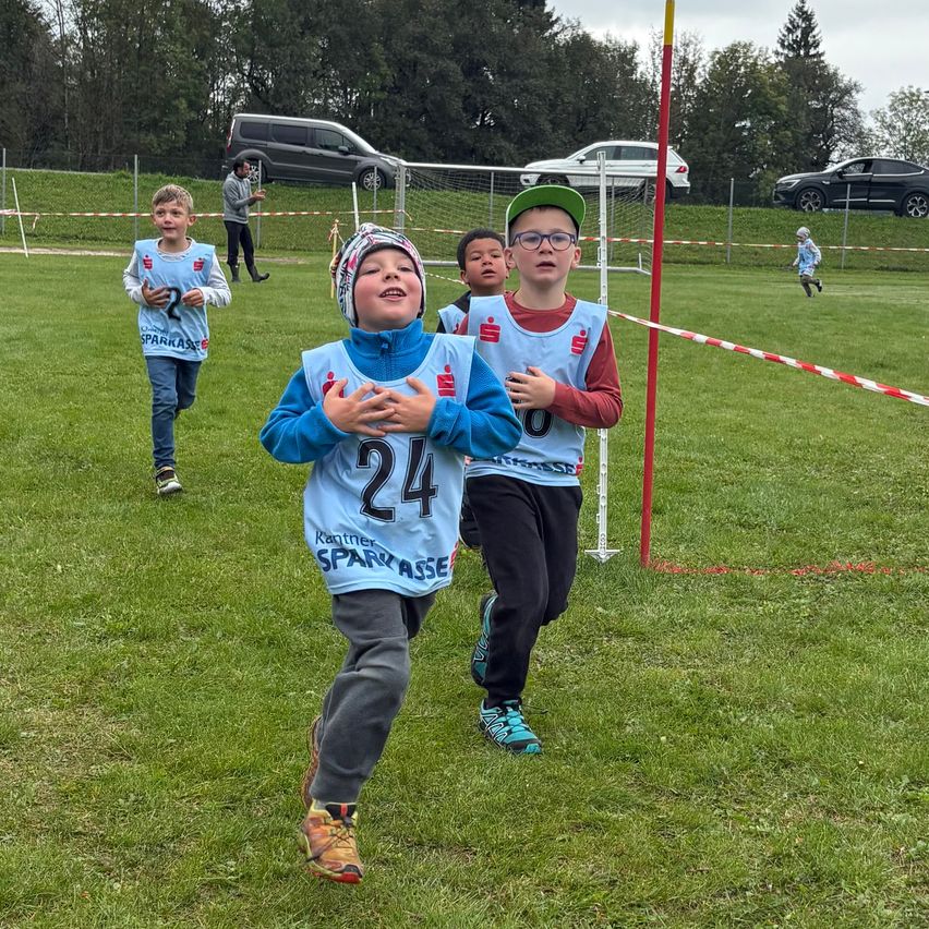Kinder in Trikots mit Nummern laufen auf einem Rasenfeld. Sie nehmen an einem Rennen teil. Ein Fußballtor und rot-weiße Barrieren sind im Hintergrund.