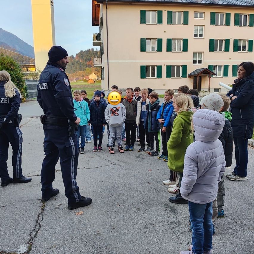 Ein Polizist spricht vor einem Gebäude mit grünen Fensterläden zu einer Reihe von Kindern, einige in Turnschuhen.