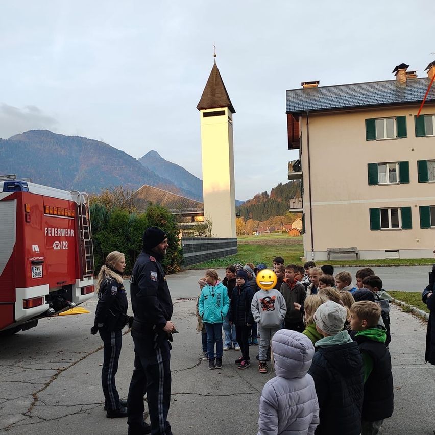 Zwei Feuerwehrleute stehen vor einer Gruppe von Kindern, in der Nähe eines Feuerwehrwagens. Sie befinden sich in einer Stadt mit Gebäuden, Bergen und einem bewölkten Himmel im Hintergrund.