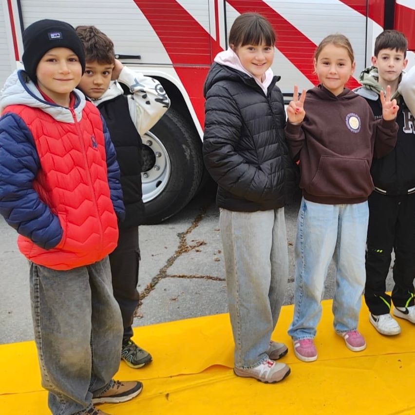 Fünf Kinder stehen vor einem Feuerwehrauto, alle in Winterkleidung. Zwei von ihnen machen Friedenszeichen.