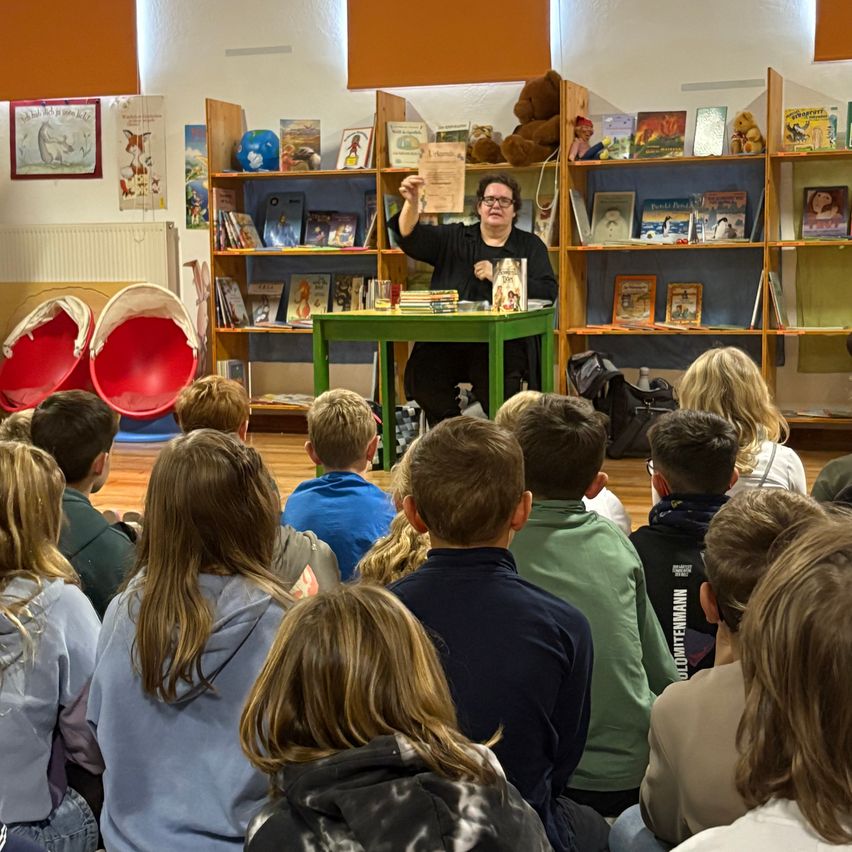 Eine erwachsene Frau liest ein Buch in einem Klassenzimmer vor einer Gruppe von Kindern, die auf dem Boden sitzen. Hinter ihr befinden sich Regale voller Bücher und Plüschtiere.