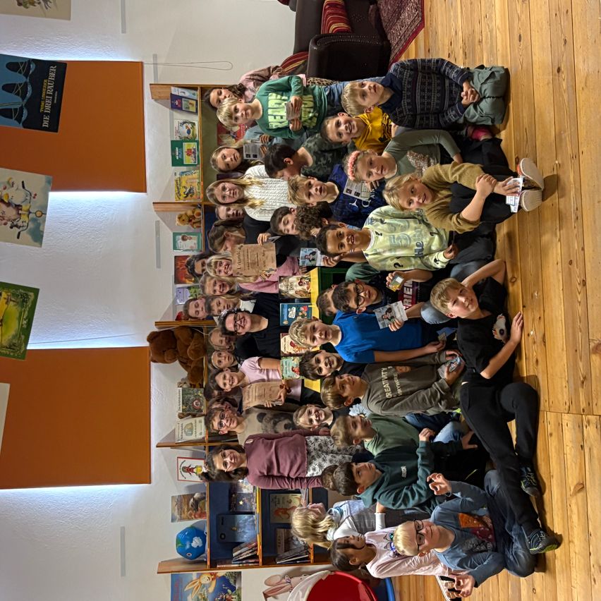 Eine Gruppe von Kindern, einige in Freizeitkleidung, sind um einen Tisch versammelt und posieren möglicherweise für ein Foto. Der Raum hat Holzböden, Regale voller Bücher und eine orangefarbene Wand.