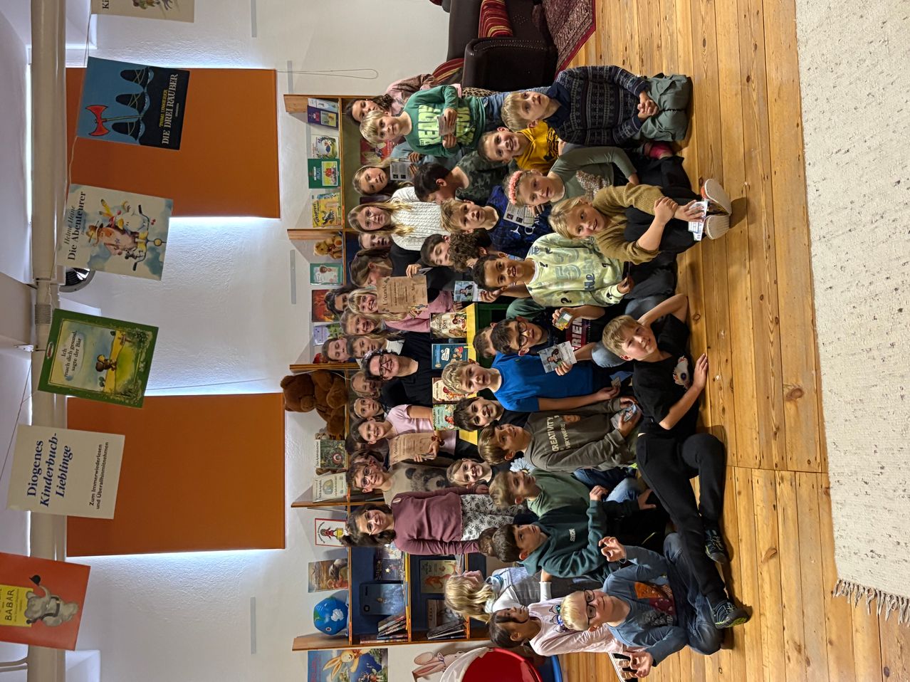 Eine Gruppe von Kindern, einige in Freizeitkleidung, sind um einen Tisch versammelt und posieren möglicherweise für ein Foto. Der Raum hat Holzböden, Regale voller Bücher und eine orangefarbene Wand.
