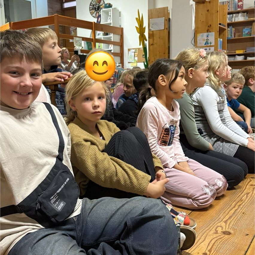 Eine Gruppe von Kindern sitzt auf dem Boden in einem Raum, einige lächeln. Ein Junge vorn trägt eine Umhängetasche und ein Mädchen neben ihm trägt eine hellpinke Jacke.
