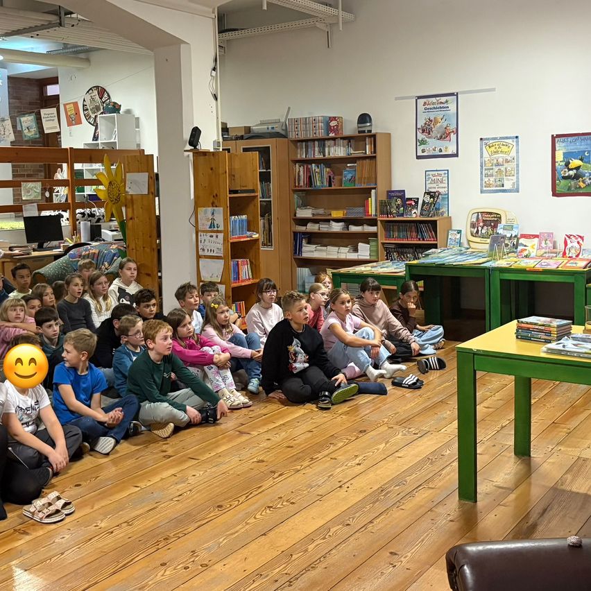Ein Klassenzimmer voller Kinder, die auf dem Boden sitzen. Regale mit Büchern und Postern schmücken die Wände. Im Hintergrund befinden sich zwei grüne Schreibtische mit Büchern.