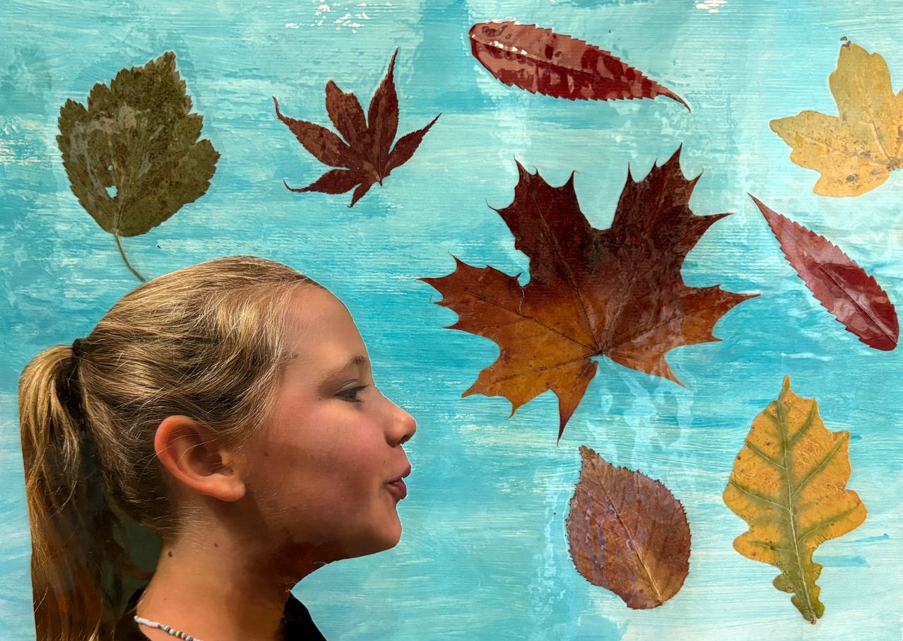 Ein junges Mädchen mit blonden Haaren schaut an einem Gemälde von Herbstblättern auf einem blauen Hintergrund nach oben.