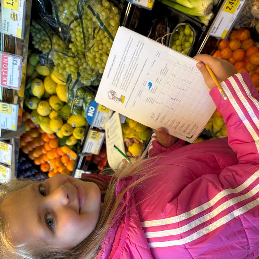 Ein junges Mädchen in einer rosa Jacke lächelt, während es einen Bleistift und ein Papier mit einer Checkliste in einem mit Obst gefüllten Lebensmittelgeschäft hält.