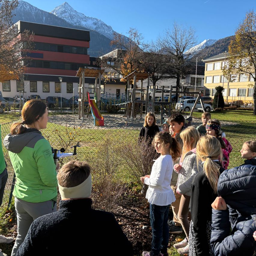 Eine Gruppe von Kindern und Erwachsenen steht in einem Park mit einem Spielplatz und Bergen im Hintergrund. Die Frau vorne trägt eine grüne Jacke und einen Kopfband.