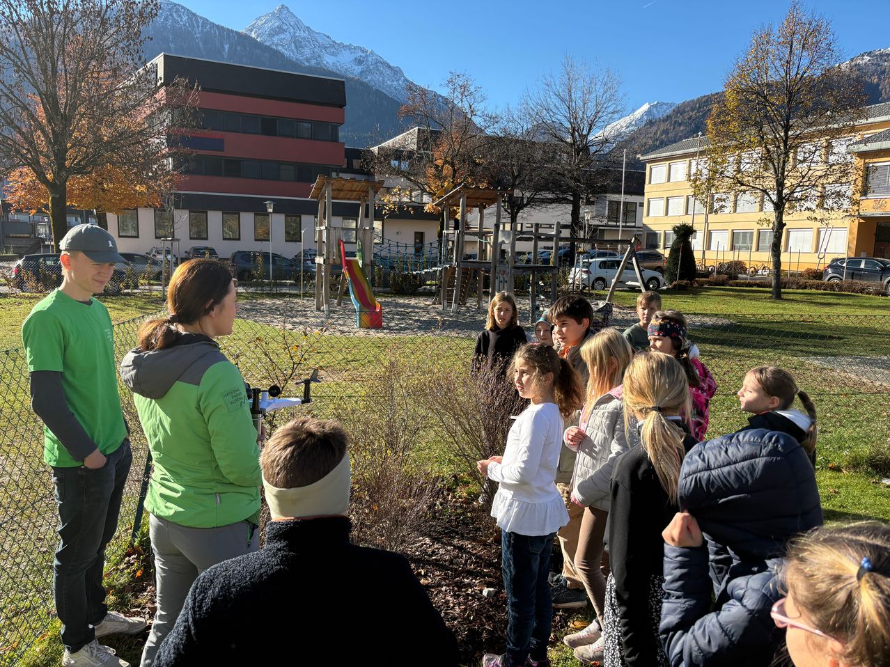 Eine Gruppe von Kindern und Erwachsenen steht in einem Park mit einem Spielplatz und Bergen im Hintergrund. Die Frau vorne trägt eine grüne Jacke und einen Kopfband.