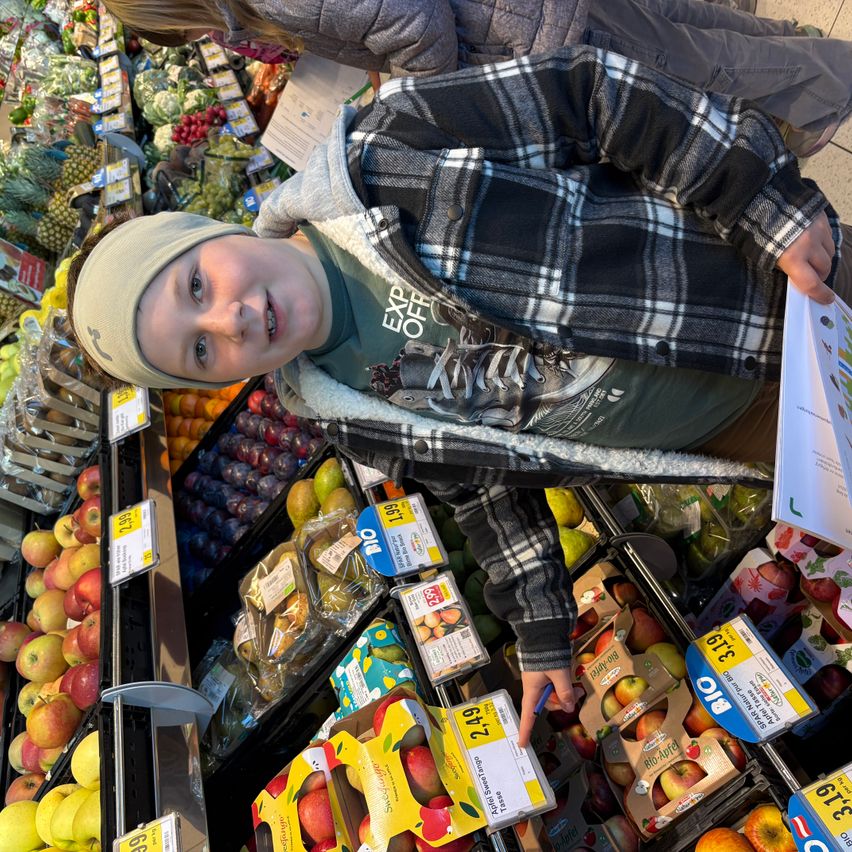 Ein junger Junge in einer karierten Jacke und Mütze steht in einem Supermarkt und hält ein Papier. Er schaut auf ein Regal mit Äpfeln mit Preisschildern.