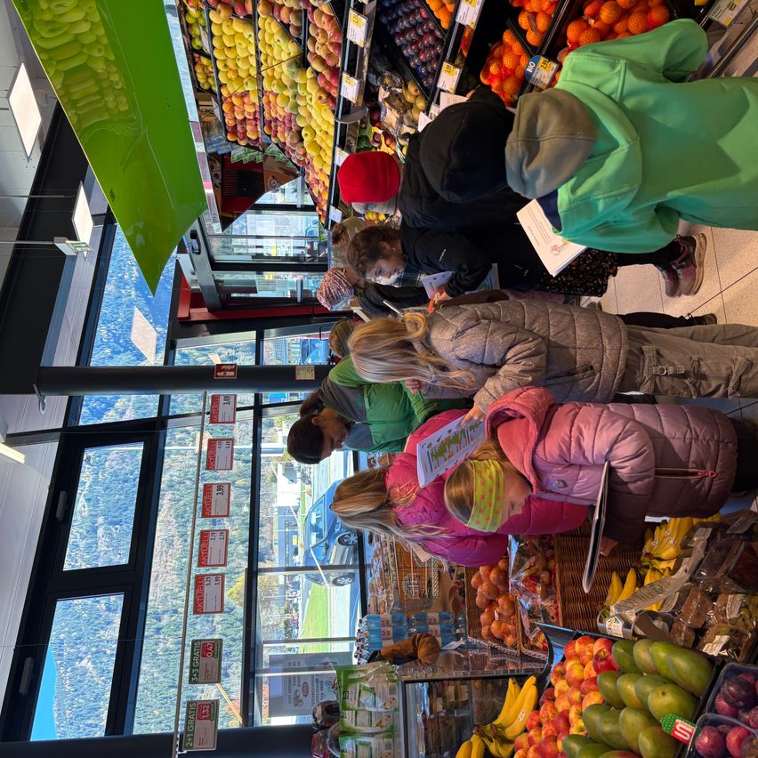 Eine Gruppe von Menschen, wahrscheinlich eine Familie, kauft in einem Supermarkt ein. Sie stöbern durch verschiedene auf Regalen ausgestellte Früchte, darunter Äpfel, Orangen und Bananen.