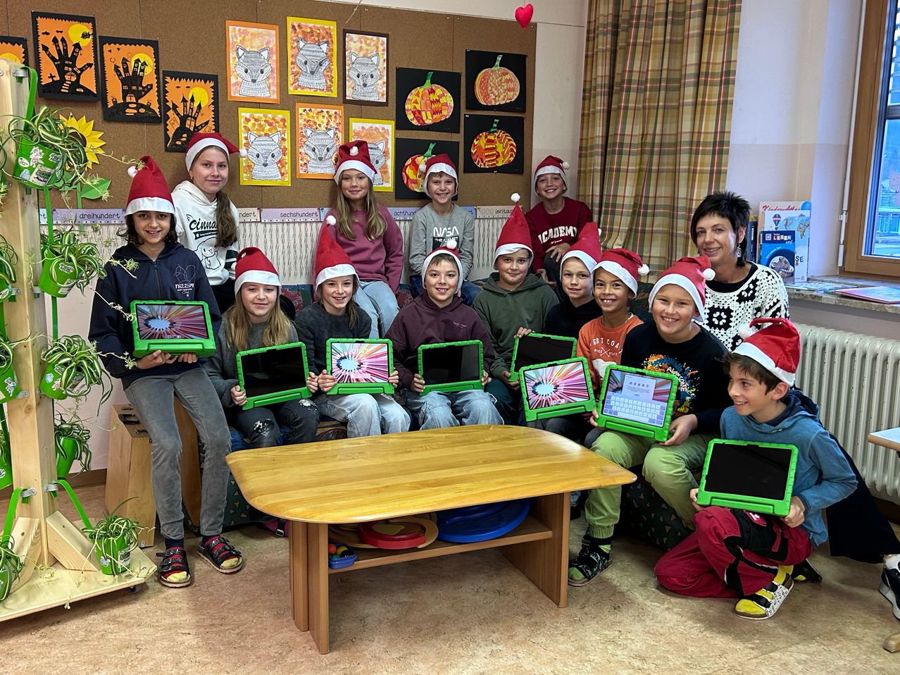 Eine Gruppe von Kindern mit Weihnachtsmützen lächelt und posiert für ein Foto, während sie Tablets halten. Sie sitzen um einen Tisch herum, auf dem verschiedene Gegenstände stehen, und es gibt Dekorationen an der Wand hinter ihnen.