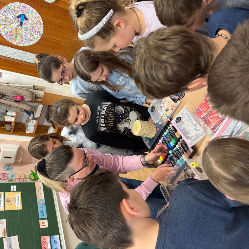 Eine Gruppe von Kindern und Erwachsenen versammelt sich um einen Tisch mit verschiedenen Kunstmaterialien. Sie sind in einer künstlerischen Aktivität vertieft. Links ist eine Tafel mit Text und Zahlen zu sehen.