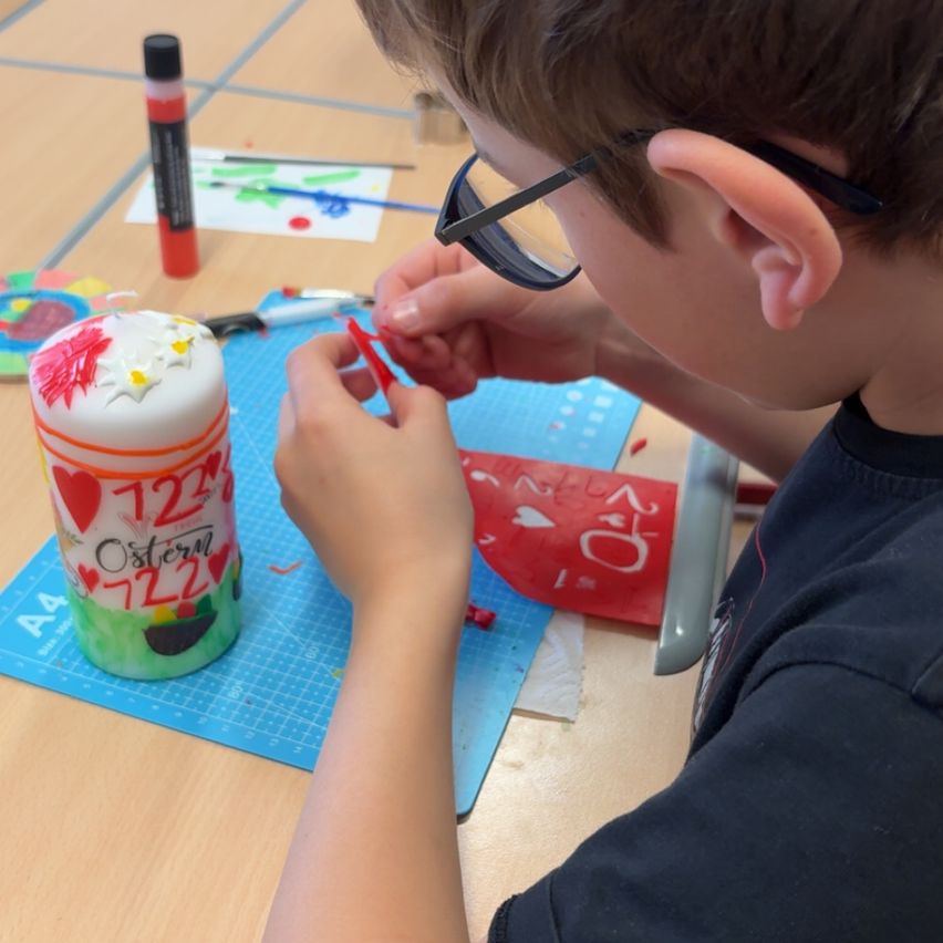 Ein Junge mit Brille verziert einen Kuchen mit rotem Papier. Er sitzt auf einem Stuhl an einem Tisch mit blauen Papieren.
