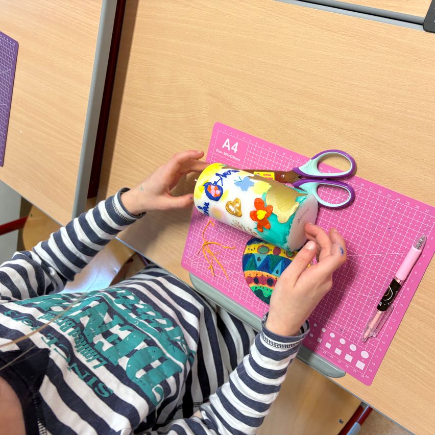 Ein Kind sitzt an einem Schreibtisch und hält einen Becher mit einem bunten Design in der Hand, wobei es eine Schere auf einem rosa Untersetzer benutzt. Der Tisch hat eine Holzoberfläche mit einem Rastermuster.