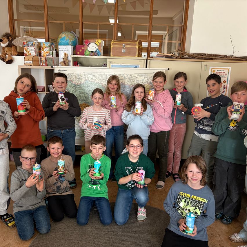 Eine Gruppe von Kindern posiert für ein Foto und hält dekorierte Becher hoch, in einem Raum mit Regalen und einer Karte an der Wand.