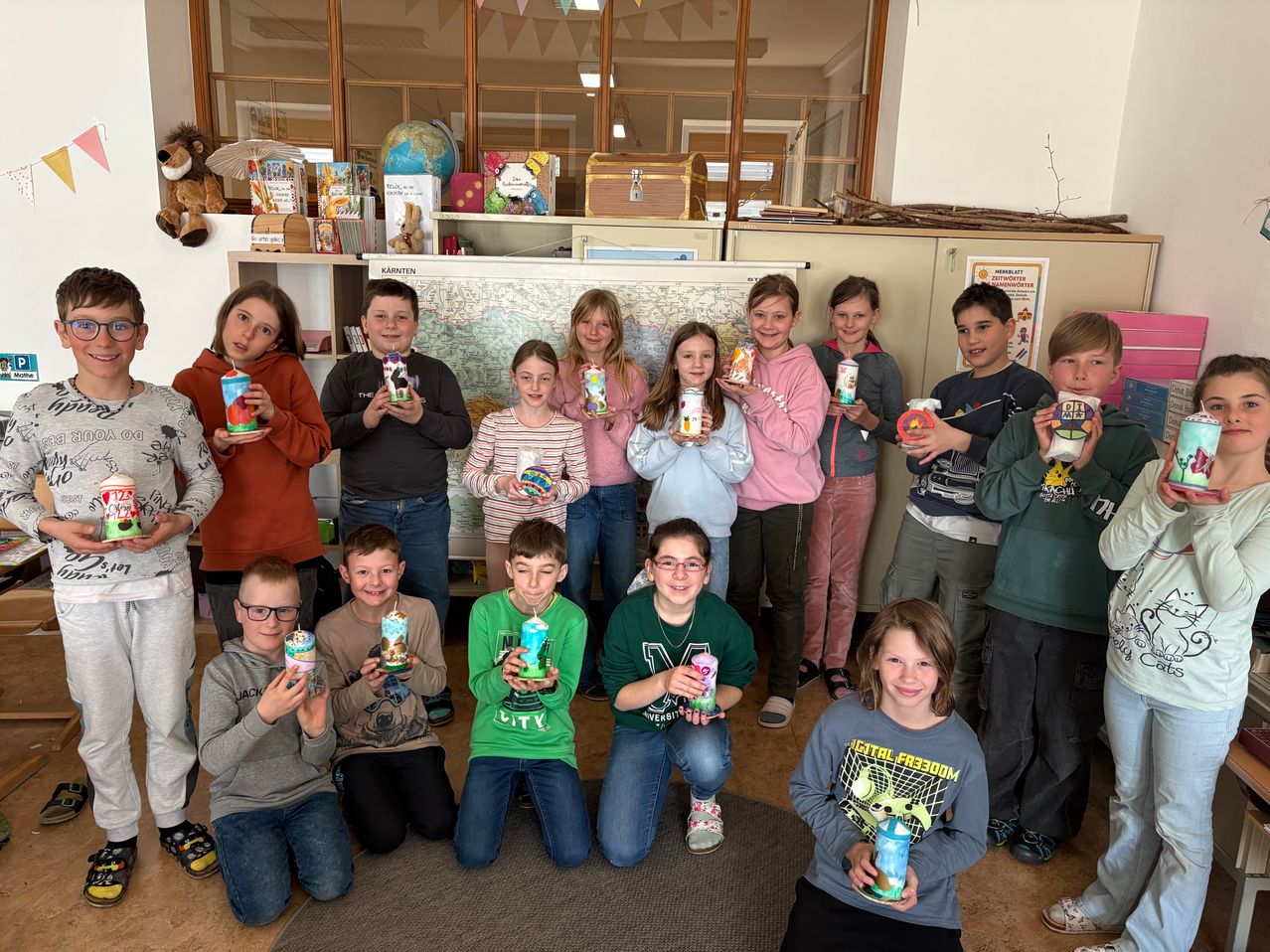 Eine Gruppe von Kindern posiert für ein Foto und hält dekorierte Becher hoch, in einem Raum mit Regalen und einer Karte an der Wand.