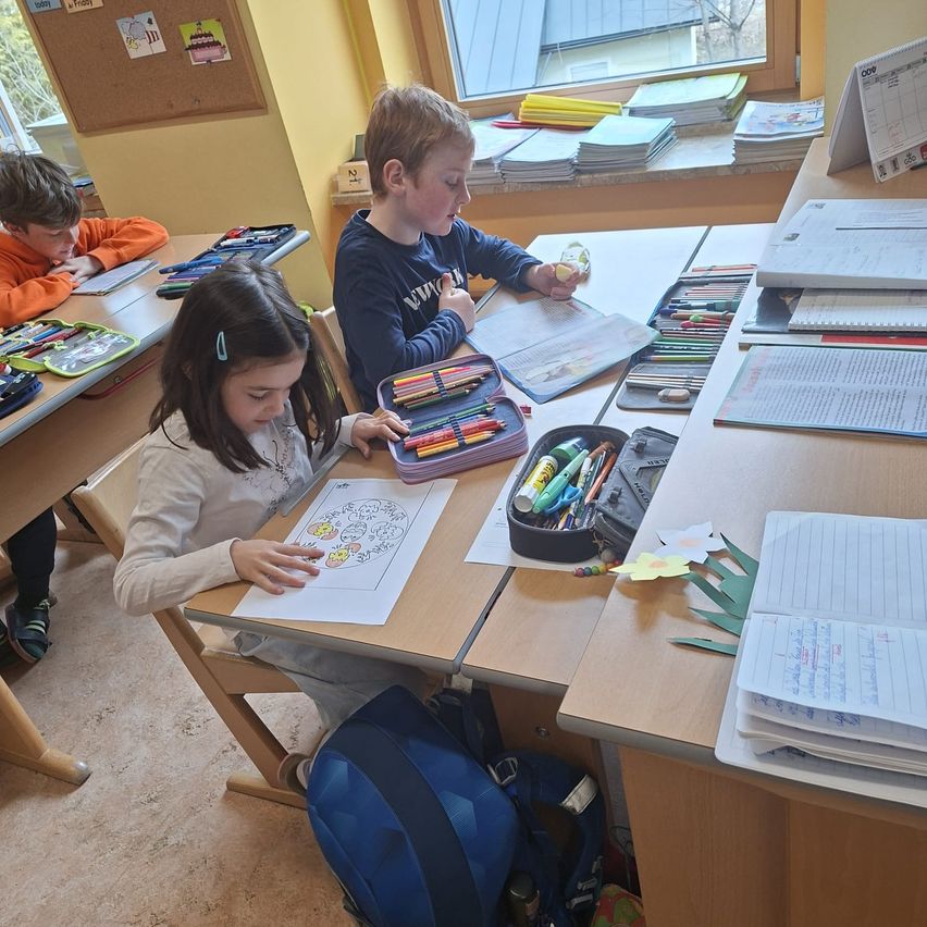 Drei Kinder sitzen an Schreibtischen in einem Klassenzimmer. Das Mädchen vorne hält ein Malbuch. Der Junge neben ihr schaut in einen Malstifte-Behälter. Ein anderer Junge sitzt hinten.