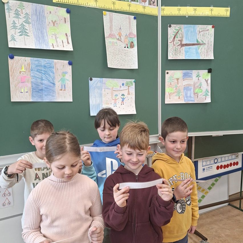 Fünf Kinder stehen vor einer Tafel mit Zeichnungen und halten Papiere in der Hand. Sie scheinen an einer Aktivität beteiligt zu sein. Hinter ihnen ist die Tafel mit verschiedenen Zeichnungen beheftet.