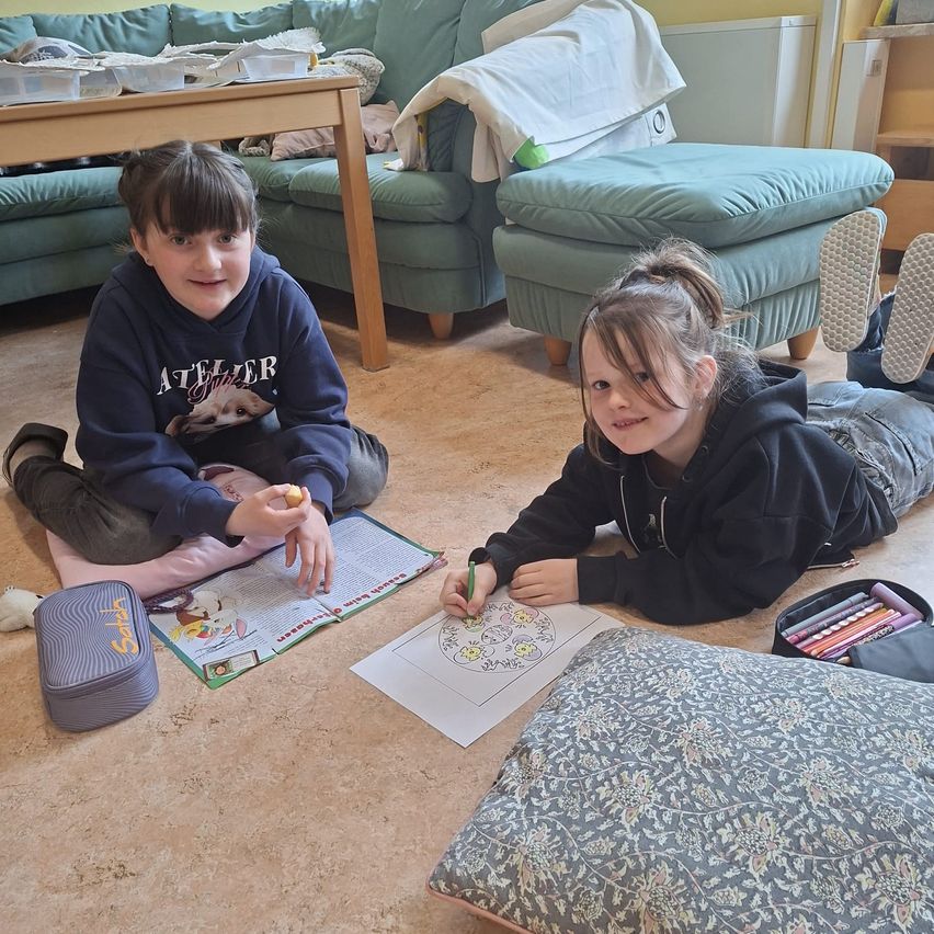 Zwei junge Mädchen sitzen auf dem Boden und zeichnen ein Bild. Eine hält ein Malbuch und die andere zeichnet auf Papier. Sie haben Malstifte und eine Federmappe in der Nähe. Dahinter steht eine Couch mit Kissen und ein Holztisch mit verschiedenen Gegenständen.