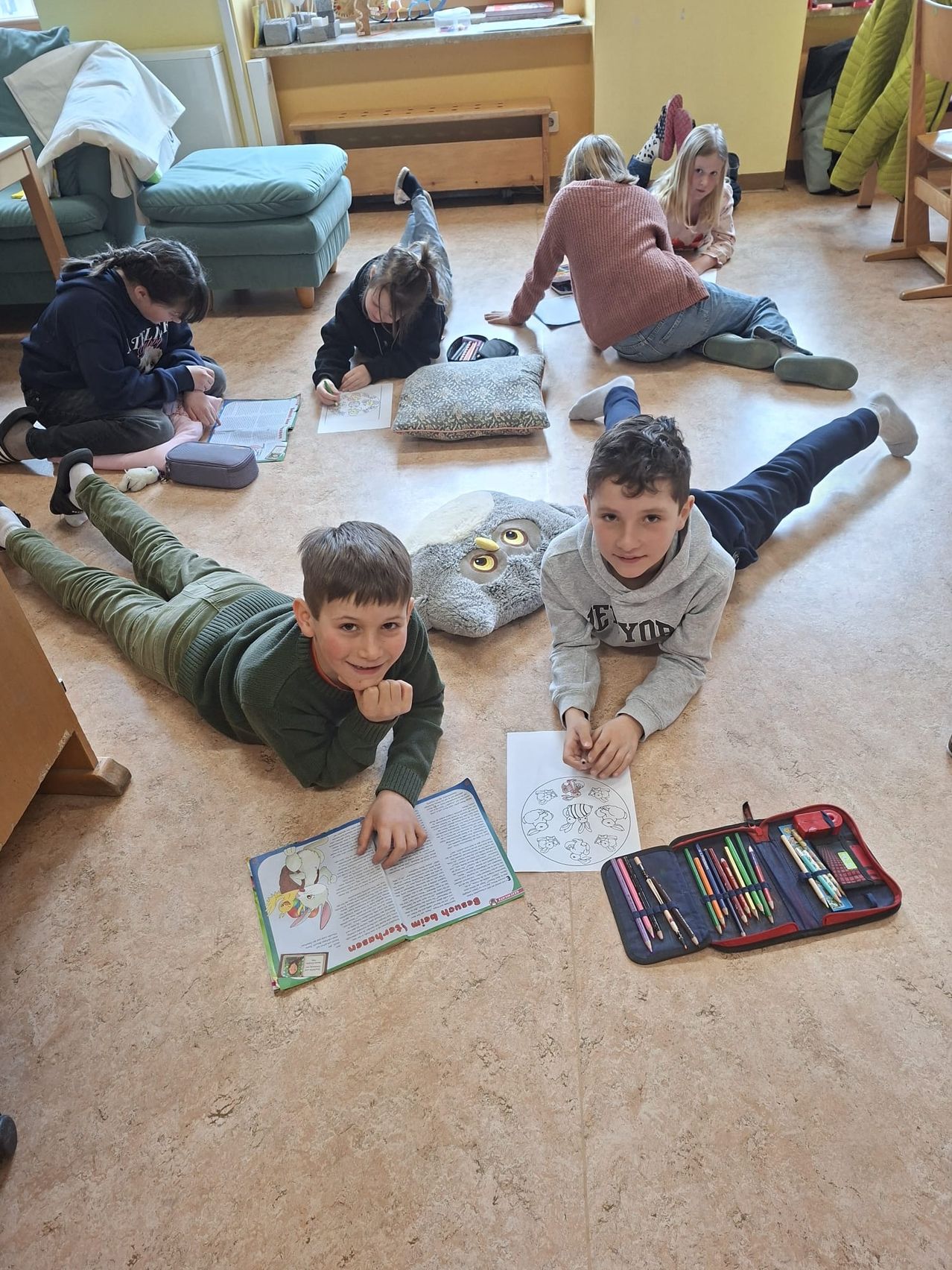 Kinder in einem Klassenzimmer sitzen und liegen auf dem Boden und machen ein Kunstprojekt mit Malbüchern, Stiften und einem Eulenkissen.