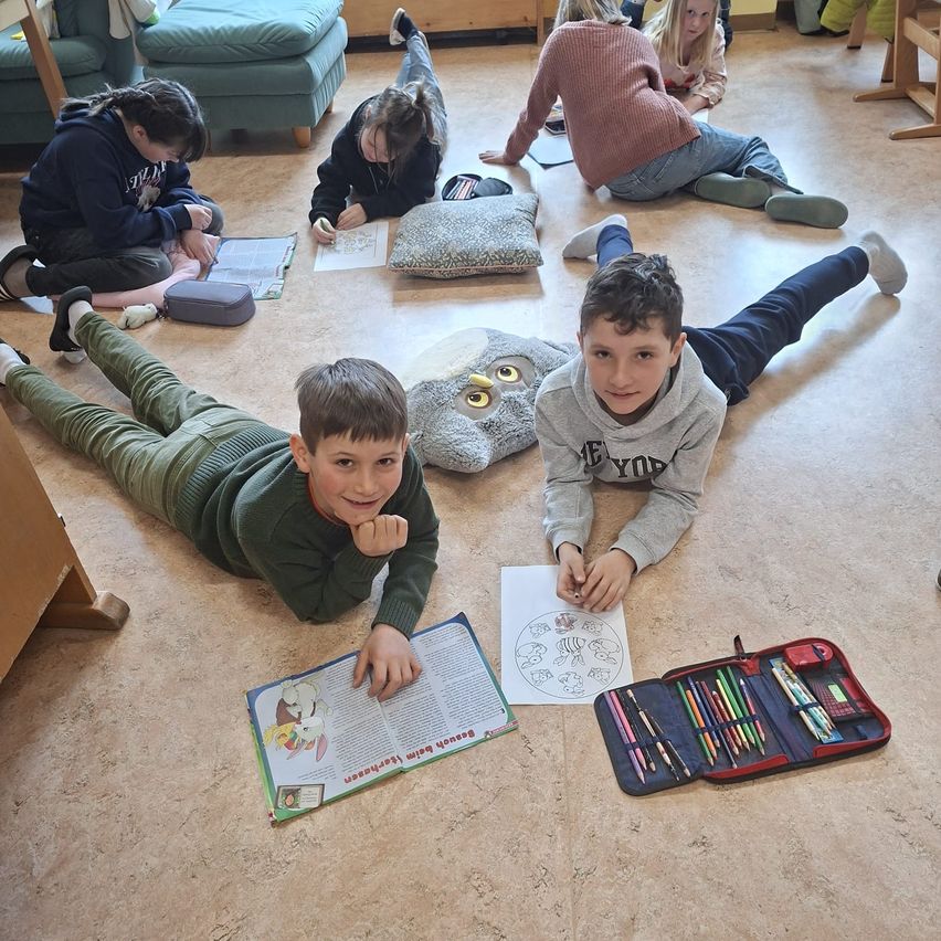 Kinder in einem Klassenzimmer sitzen und liegen auf dem Boden und machen ein Kunstprojekt mit Malbüchern, Stiften und einem Eulenkissen.