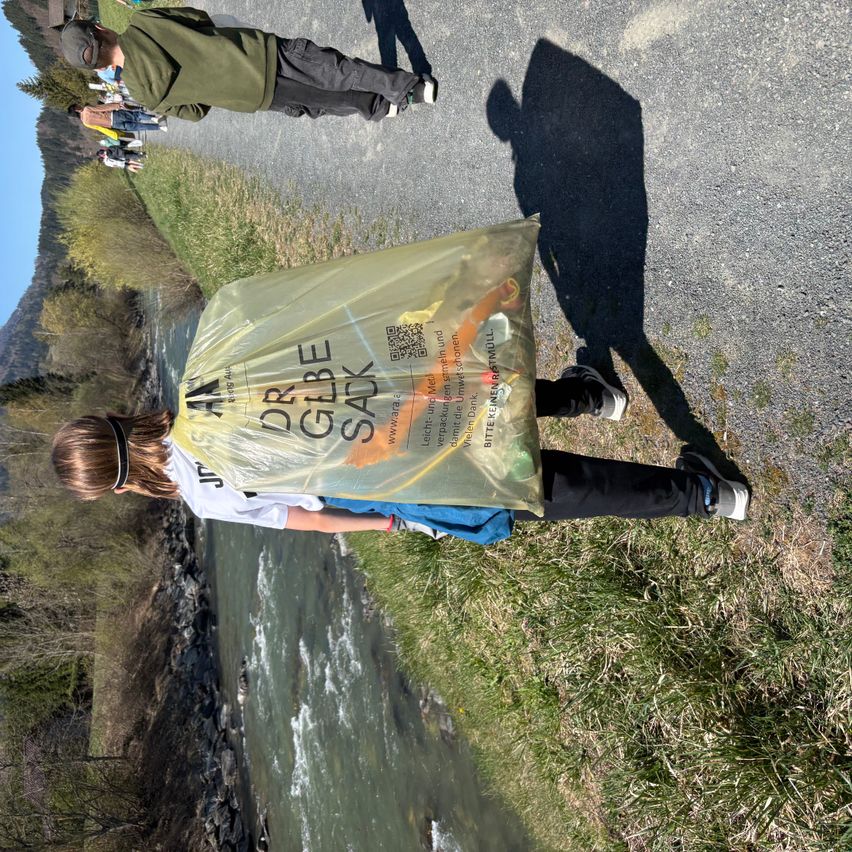 Eine Person, die einen mit einem QR-Code versehenen Dr. Globke Sack trägt, steht am Fluss, während eine weitere Person auf der Straße hinter ihr geht.