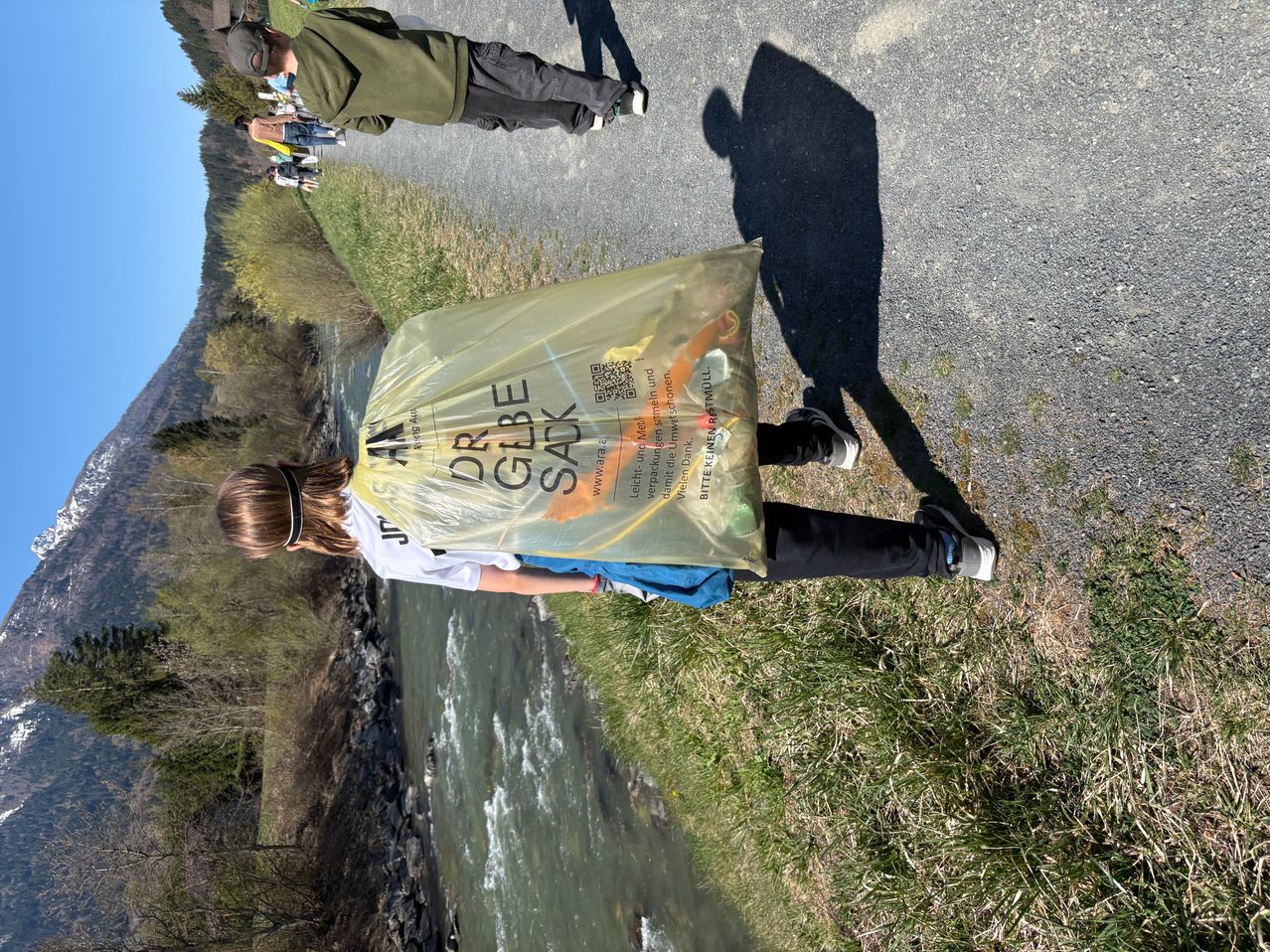 Eine Person, die einen mit einem QR-Code versehenen Dr. Globke Sack trägt, steht am Fluss, während eine weitere Person auf der Straße hinter ihr geht.