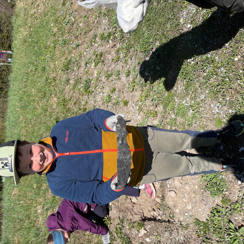 Ein Kind hält einen grauen Felsen, lächelt und posiert für ein Foto auf einem Grasboden mit zwei anderen Kindern in der Nähe.
