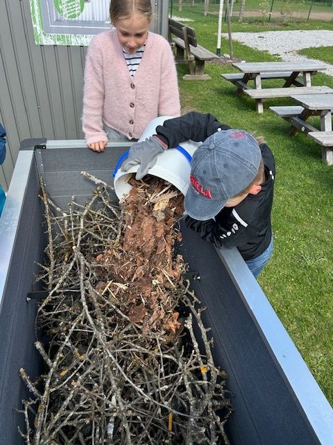 Ein Junge schüttet einen Eimer in einen Behälter, der mit Stöcken und Blättern gefüllt ist. Eine Mädchen steht daneben und beobachtet. Dahinter steht eine Holzbank und ein Schild.