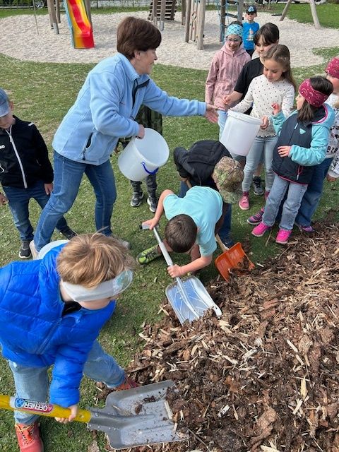 Kinder in Winterjacken räumen im Park Laub unter Aufsicht von Erwachsenen auf. Einige tragen Eimer. Im Hintergrund sind ein Rutsch und Spielgeräte zu sehen.