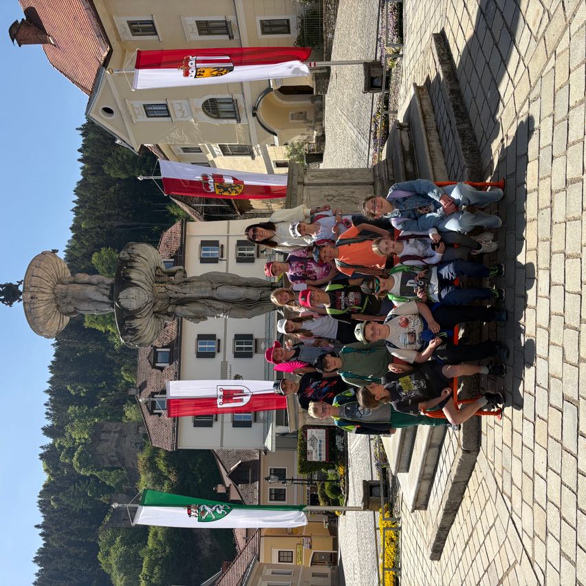 Eine Gruppe von Kindern posiert für ein Foto vor einem Brunnen, umgeben von Gebäuden mit Fahnen und einer Statue.