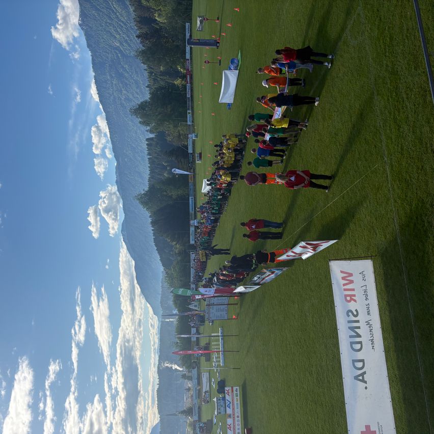 Eine Gruppe von Menschen, wahrscheinlich Teilnehmer, steht auf einem grünen Feld mit Bannern und Flaggen. Ein Gebirgszug ist im Hintergrund sichtbar.