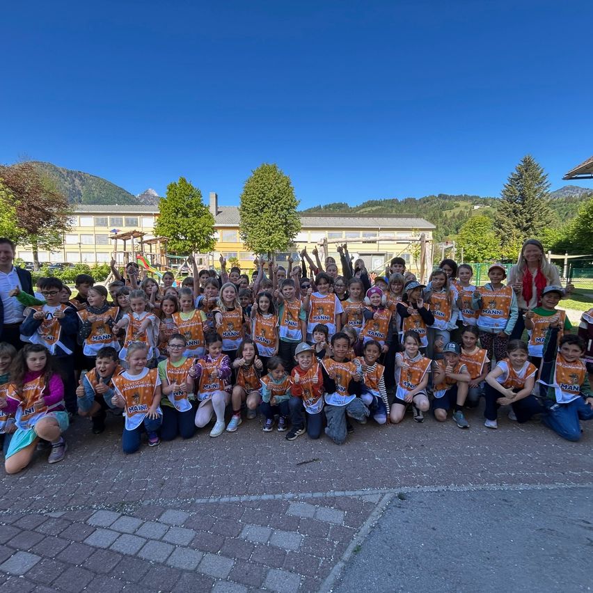 Eine Gruppe von Kindern und Erwachsenen posiert für ein Foto im Freien. Sie tragen orangefarbene T-Shirts mit dem Wort 'MANN'. Dahinter steht ein Spielplatz und Gebäude. Bäume und Berge sind im Hintergrund.