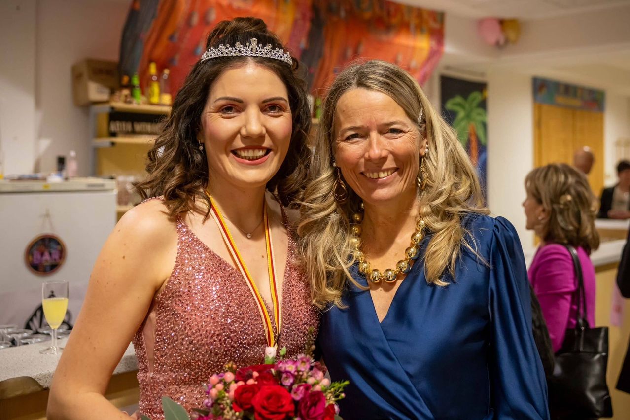 Zwei lächelnde Frauen, eine in einem rosa Paillettenkleid mit Krone, die andere in einem blauen Kleid, Blumenstrauß in der Hand, in einem dekorierten Raum.