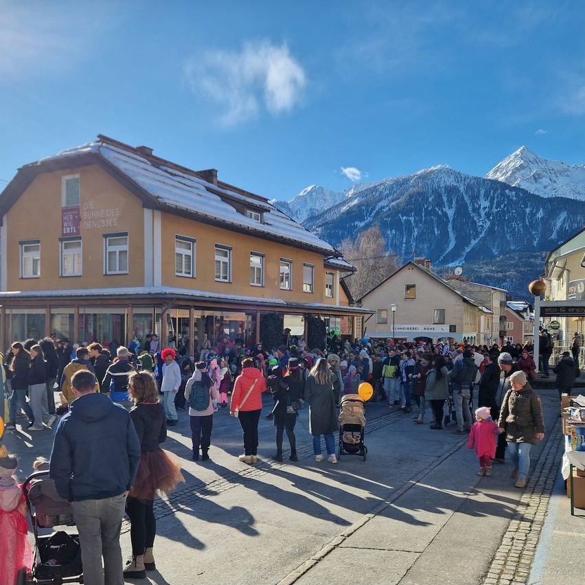 Eine Menschenmenge geht auf einer Straße vor einem Gebäude mit schneebedecktem Dach. Viele Menschen tragen Kostüme. Ein Kinderwagen steht auf der Straße. Eine Person hält einen gelben Luftballon. Berge sind in der Ferne.