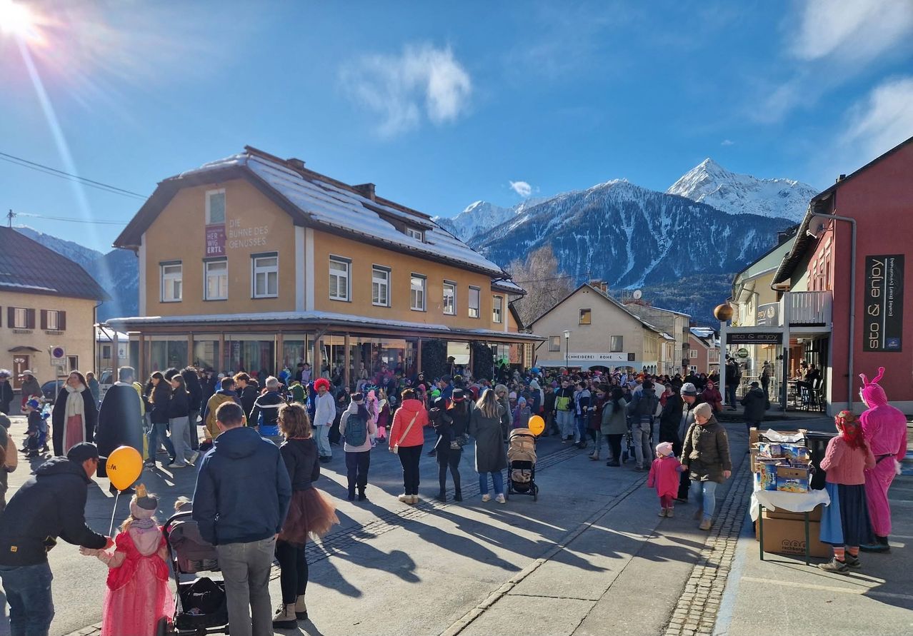 Eine Menschenmenge geht auf einer Straße vor einem Gebäude mit schneebedecktem Dach. Viele Menschen tragen Kostüme. Ein Kinderwagen steht auf der Straße. Eine Person hält einen gelben Luftballon. Berge sind in der Ferne.