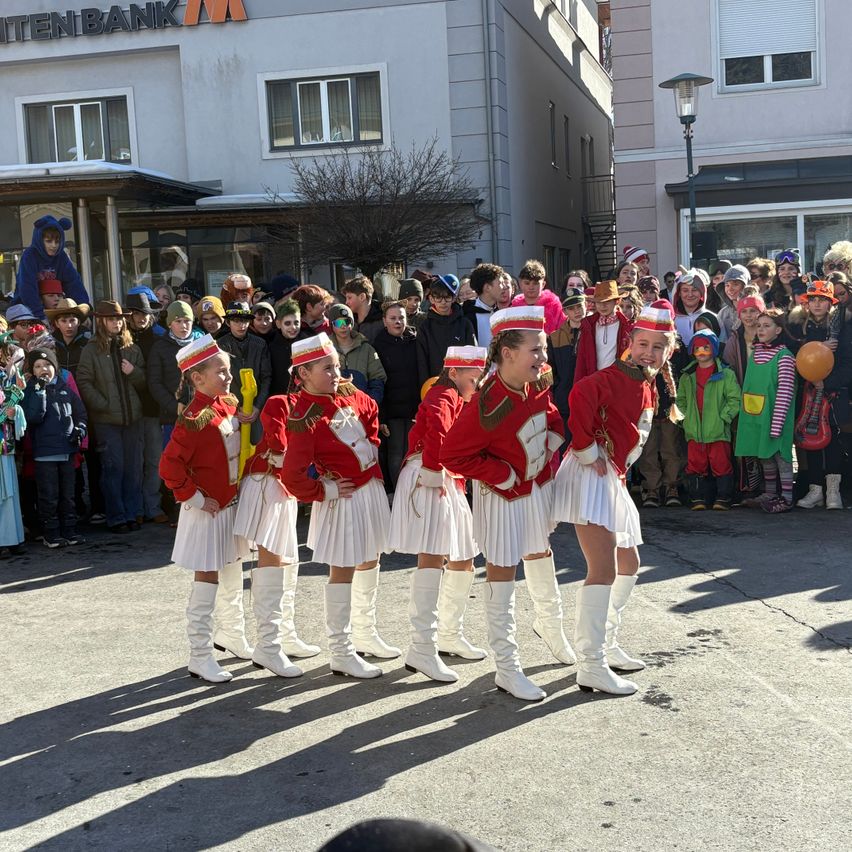 Eine Gruppe junger Menschen in roten und weißen Uniformen mit weißen Stiefeln steht in einer Reihe auf der Straße und tritt vor einer Zuschauermenge auf.