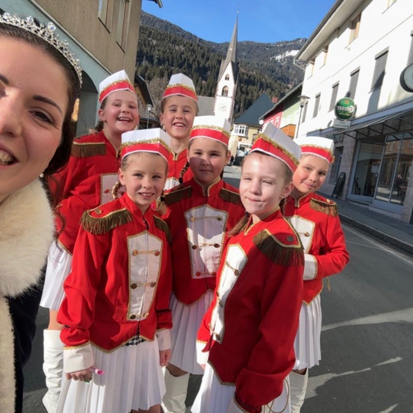 Eine Gruppe von Kindern in roten Uniformen, die an einem sonnigen Tag für ein Foto lächeln, mit einem Kirchturm und Bergen im Hintergrund.