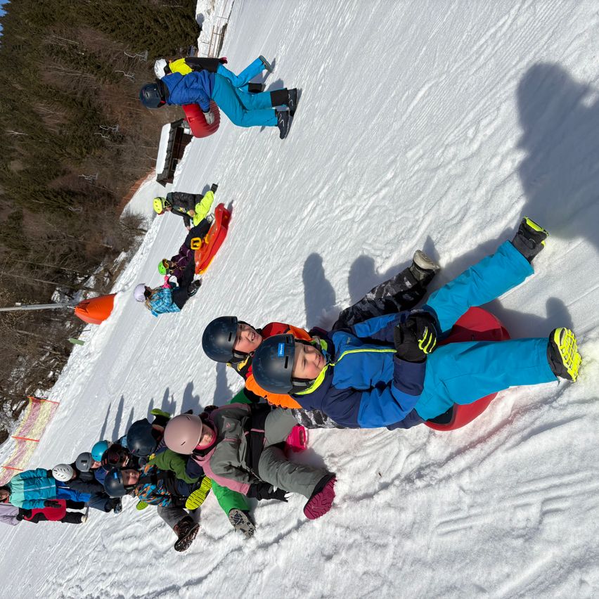 Mehrere Kinder rodeln einen schneebedeckten Hang hinunter, einige zu zweit, andere allein. Alle tragen Winterkleidung und Helme. Bäume und ein roter Verkehrskegel sind im Hintergrund zu sehen.
